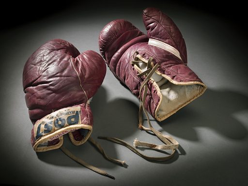 Dark red brown training boxing gloves worn by Cassius Clay (Muhammad Ali) and signed in Louisville, as indicated by the penned note inside the left glove near the wrist. Gloves are edged in faded yellow fabric and tied with dark, faded strings. Stitching throughout the glove is in white thread. On the outside fronts of both gloves below the wrist are manufacturers labels. [POST / MANUFACTURING CO. / NEW YORK]. Inside the left glove, near the wrist, is a note in faded red ink. [SIGNED BY / CASSIUS CLAY/ Louisville / 1960]. Inside the right glove, near the wrist, is a note in blue pen [From / Cassius Clay / 1960]. Gloves are well worn and wrinkled. The right glove has dark discolourations near the top of the glove on the right side. Date: 1960s. Record ID: nmaahc_2012.173.3ab.