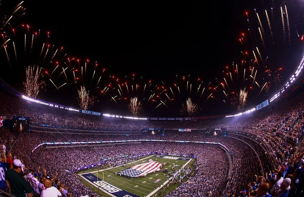 The biggest stage in America is illuminated with red, white, and blue fireworks. In the center of the field, people hold up a large  and beautiful American flag. 